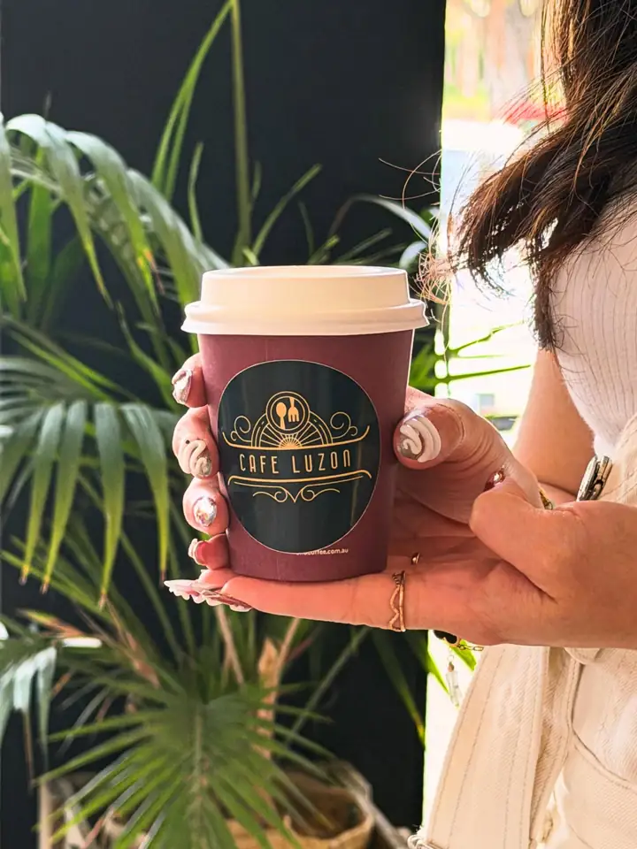 A Café Luzon takeaway coffee cup held up in front of palm leaves